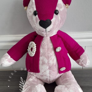 May include: A pink and magenta teddy bear wearing a magenta cardigan with pearl and rhinestone embellishments. The bear has a black heart-shaped nose and black eyes. The bear's paws have the words "Fern Dreams of Fairies" and "Mum" on one paw and "Goodnight & Sleep Well" on the other.