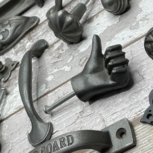 May include: A selection of antique-style cabinet hardware. The image shows a variety of knobs, pulls, and handles in a dark grey finish. A unique piece is a hand-shaped knob giving a thumbs-up.