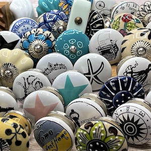 May include: Assortment of ceramic drawer knobs with various designs, including a starfish, an octopus, a compass, a typewriter, and a floral pattern. The knobs are white with colourful accents and have a silver finish.