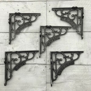 May include: Five black cast iron shelf brackets with ornate designs. Each bracket has a different London station name: Paddington, St Pancras, Victoria, Euston, and Kings Cross.