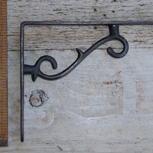 May include: Black metal shelf bracket with a decorative scroll design. The bracket is approximately 15 cm long.