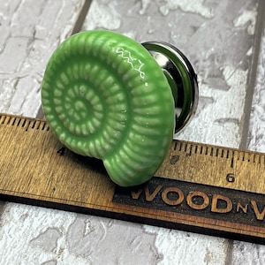 May include: Green ceramic cabinet knob shaped like a spiral seashell. The knob is approximately 7.5 cm long and has a silver metal base.