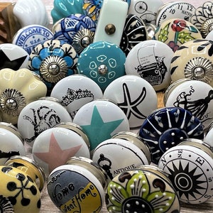 May include: Assortment of ceramic drawer knobs with various designs, including a starfish, an octopus, a compass, a typewriter, and a floral pattern. The knobs are white with colourful accents and have a silver finish.