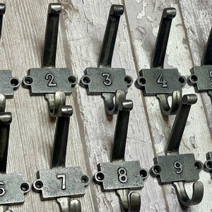 May include: A collection of ten antique-style metal coat hooks, each numbered from one to ten. The hooks are a dark grey colour and mounted on a light-coloured, distressed wooden surface. Each hook has a rectangular plate with a number.