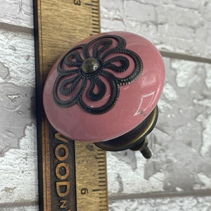 May include: A pink ceramic cabinet knob with a black metal flower design. The knob has a brass screw.