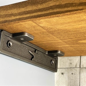 May include: A close-up of a dark grey metal shelf bracket with a decorative design. The bracket is attached to a wooden shelf.