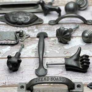 May include: A collection of 16 black and silver metal cabinet knobs and pulls, including a bear, a hand giving a thumbs up, and a pull that says "Bread Board".