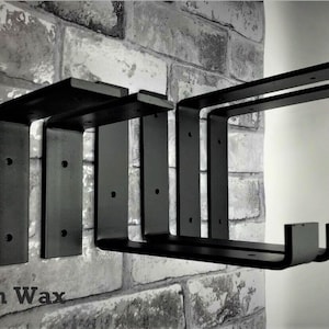 May include: Four black metal shelf brackets mounted on a brick wall. The brackets are L-shaped with a flat top and a curved bottom. The brackets are all the same size and are arranged in a row. The text "Wood n Wax" is visible in the bottom left corner of the image.