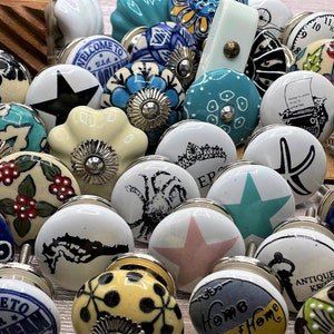May include: Assortment of ceramic knobs with various designs and colours, including floral, nautical, and geometric patterns. Some knobs feature text such as "Welcome to" and "Home".