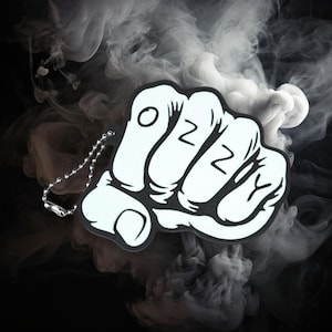 Op de afbeelding: Een zwart-witte vuistvormige sleutelhanger met de letters "OZZY" erop gedrukt. De sleutelhanger heeft een zilveren ketting en staat tegen een rokerige achtergrond.