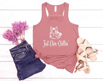 May include: A pink tank top with a white graphic of a chinchilla and the text "Just Chin-Chillin'" on a white wooden background.