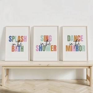 May include: Three framed prints with colorful text. The first print says "SPLASH in the BATH", the second print says "SING in the SHOWER", and the third print says "DANCE in the MIRROR". The prints are hanging on a white wall above a wooden bench.