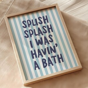 May include: A framed print with the text "SPLISH SPLASH I WAS HAVIN' A BATH" in navy blue on a light blue and white striped background. The frame is made of light-coloured wood.