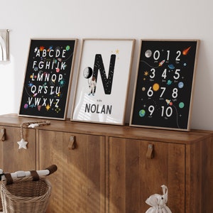 May include: Three black framed prints with a space theme. The first print features the alphabet with planets and stars. The second print features a large letter N with a spaceman and the name Nolan. The third print features numbers 0-10 with planets and stars.