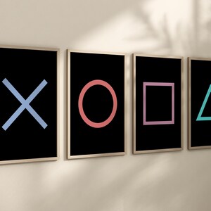 May include: Four framed prints on a white wall, each featuring a different coloured symbol from a video game controller: a blue X, a red circle, a pink square, and a turquoise triangle.