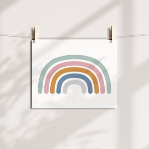 May include: A square art print featuring a minimalist rainbow design. The rainbow has layers of pastel colors: light blue, pink, gold, dark blue, and gray. The print is hanging from a string with wooden clips against a white wall.