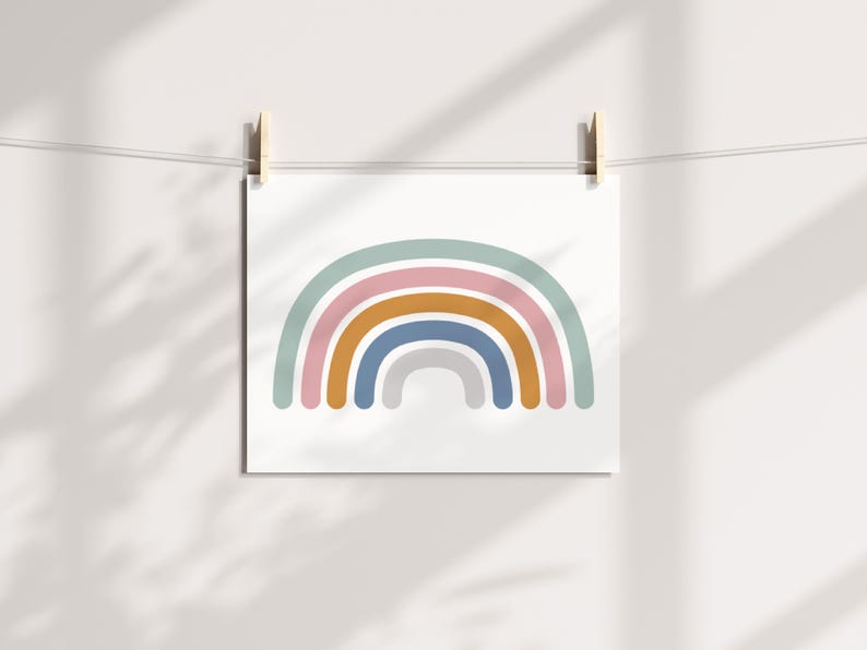 May include: A square art print featuring a minimalist rainbow design. The rainbow has layers of pastel colors: light blue, pink, gold, dark blue, and gray. The print is hanging from a string with wooden clips against a white wall.
