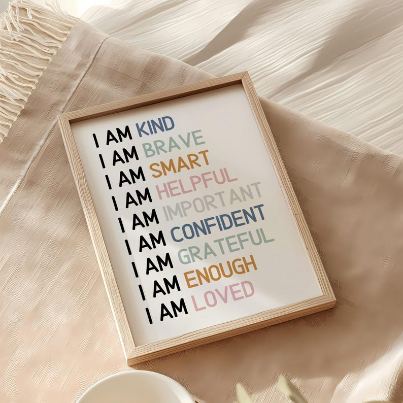May include: A framed print with the words "I AM KIND, BRAVE, SMART, HELPFUL, IMPORTANT, CONFIDENT, GRATEFUL, ENOUGH, LOVED" in various colors. The frame is light brown, and the background is white. The print is on a beige surface.