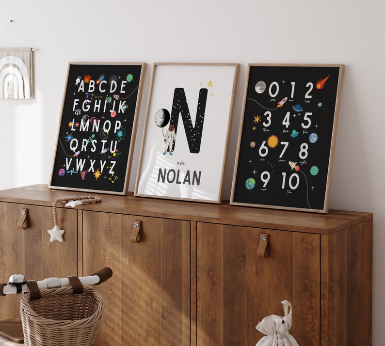 May include: Three black framed prints with a space theme. The first print features the alphabet with planets and stars. The second print features a large letter N with a spaceman and the name Nolan. The third print features numbers 0-10 with planets and stars.