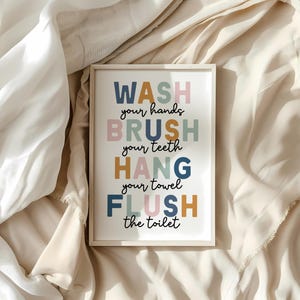 May include: A framed bathroom art print with the words "WASH your hands, BRUSH your teeth, HANG your towel, FLUSH the toilet" in colorful block letters. The print is set against a neutral background.