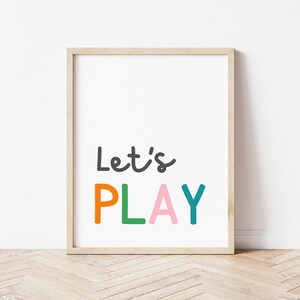 Puede incluir: Una impresi&oacute;n enmarcada con el texto "Let's PLAY" en letras de colores. Las letras son naranja, verde, rosa y turquesa. El marco es de madera marr&oacute;n claro.