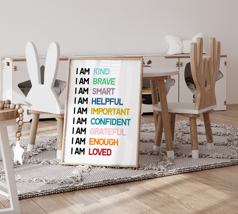 May include: A white framed poster with the text "I AM" followed by a list of positive affirmations in different colors, including "KIND", "BRAVE", "SMART", "HELPFUL", "IMPORTANT", "CONFIDENT", "GRATEFUL", "ENOUGH", and "LOVED". The poster is displayed in a children's playroom with two wooden chairs.
