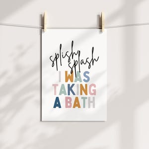 May include: A white print with the words "splishy splash" in black script above the text "I WAS TAKING A BATH" in colorful block letters. The print is held by wooden clips on a white string.