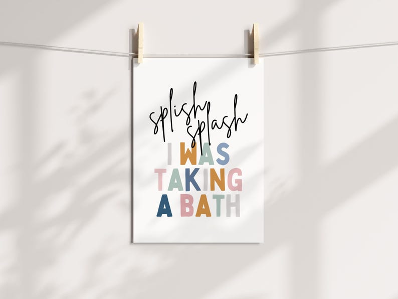 May include: A white print with the words "splishy splash" in black script above the text "I WAS TAKING A BATH" in colorful block letters. The print is held by wooden clips on a white string.