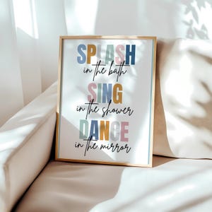 以下が含まれることがあります： カラフルな文字で「SPLASH in the bath SING in the shower DANCE in the mirror」と書かれた額装されたプリント。プリントは白い背景で、金色の額縁が付いています。