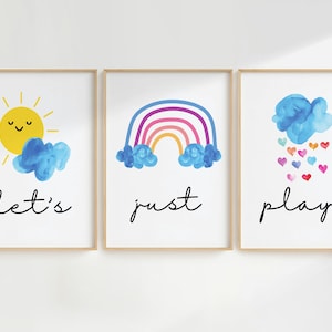 Puede incluir: Tres impresiones enmarcadas con ilustraciones de acuarela de un sol, un arcoíris y una nube con corazones. El texto "let's just play." está escrito debajo de las imágenes.