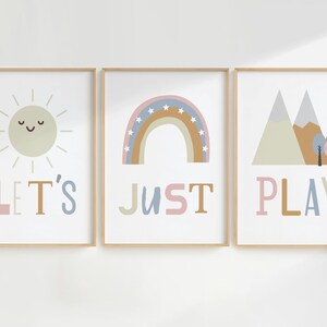 Puede incluir: Tres impresiones enmarcadas con un diseño minimalista. La primera impresión presenta un sol amarillo con una cara sonriente y el texto "LET'S". La segunda impresión presenta un arcoíris con estrellas y el texto "JUST". La tercera impresión presenta una cordillera con árboles y el texto "PLAY".