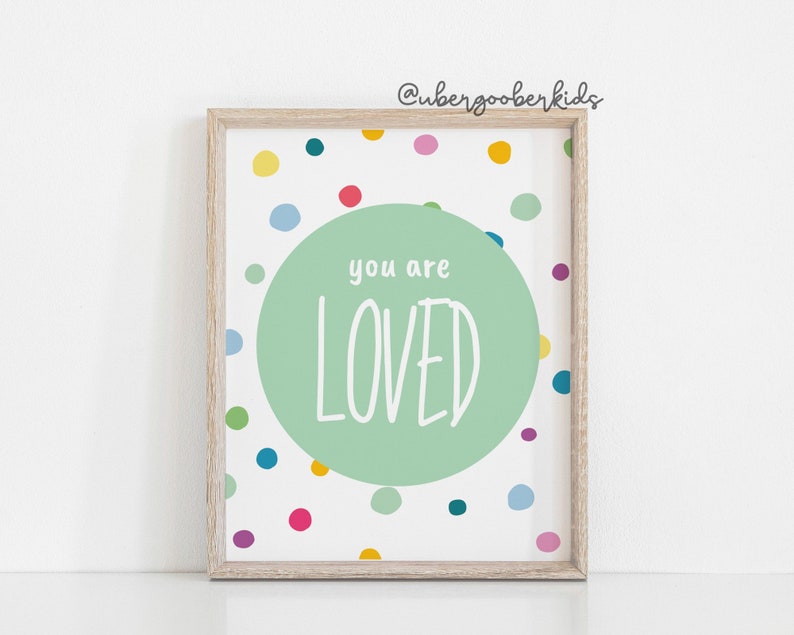 May include: A framed print with a white background and a green circle in the centre. The circle has the text "you are LOVED" in white. The background is covered in colourful dots.