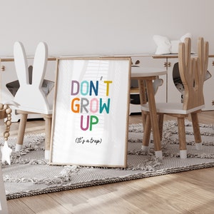 May include: A white framed print with the text "DON'T GROW UP (It's a trap)" in colorful letters. The print is hanging on a white wall in a playroom with two wooden chairs and a small table.