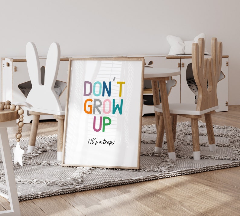 May include: A white framed print with the text "DON'T GROW UP (It's a trap)" in colorful letters. The print is hanging on a white wall in a playroom with two wooden chairs and a small table.
