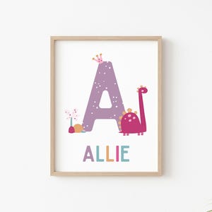 Puede incluir: Una letra A morada con lunares blancos, una corona rosa, una flor rosa y un dinosaurio rosa con manchas naranjas. El nombre Allie est&aacute; escrito debajo de la letra en letras de colores.