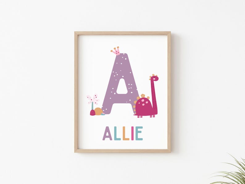 Puede incluir: Una letra A morada con lunares blancos, una corona rosa, una flor rosa y un dinosaurio rosa con manchas naranjas. El nombre Allie est&aacute; escrito debajo de la letra en letras de colores.