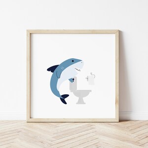 May include: A blue shark cartoon illustration with an open mouth, holding onto a white toilet with a roll of toilet paper on the holder. The image is framed in a light wood frame.