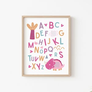 Puede incluir: Un p&oacute;ster de alfabeto colorido con un dinosaurio rosa y un cactus. Las letras est&aacute;n en diferentes colores y fuentes. El p&oacute;ster est&aacute; enmarcado en un marco de madera claro.