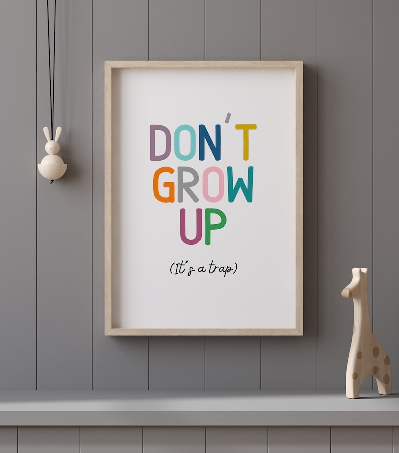 May include: A framed print with the text "DON'T GROW UP (It's a trap)" in colorful letters. The letters are in different colors, including pink, orange, green, blue, and yellow. The print is hanging on a gray wall.