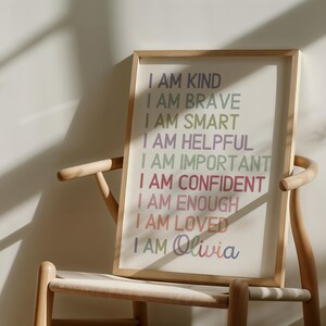 May include: A framed print with the text "I AM KIND, I AM BRAVE, I AM SMART, I AM HELPFUL, I AM IMPORTANT, I AM CONFIDENT, I AM ENOUGH, I AM LOVED, I AM Olivia" in rainbow colors on a white background.