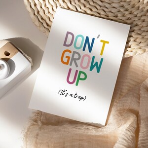 May include: A white card with the text "DON'T GROW UP (it's a trap)" in colorful letters. The card is sitting on a woven straw mat and a light brown fabric.