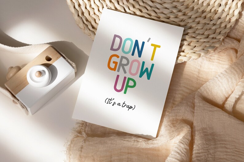 May include: A white card with the text "DON'T GROW UP (it's a trap)" in colorful letters. The card is sitting on a woven straw mat and a light brown fabric.