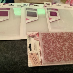 May include: Three clear plastic embossing folders in white packaging with the text "3D Embossing Folders" and "Gemini" printed on the front. The folders are for use with a die-cutting machine. A red and white patterned embossing folder is also shown in the image.