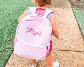 May include: A pink and white striped backpack with the name "Hazel" embroidered on the front. The backpack is being worn by a young child.
