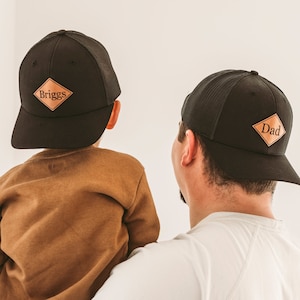 Op de afbeelding: Een zwarte mesh baseballcap met een bruin leren patch waarop "Dad" staat en een zwarte mesh baseballcap met een bruin leren patch waarop "Briggs" staat.
