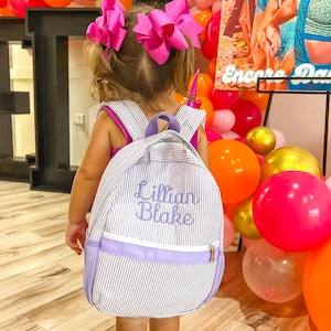May include: A child wearing a personalized backpack with the name "Lillian Blake" embroidered in blue. The backpack has a white and gray striped pattern with purple accents and a purple zipper. The child is wearing a pink top and has pink bows in their hair.