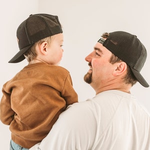 May include: A father and son wearing black baseball caps. The father is wearing a white t-shirt and the son is wearing a brown sweatshirt and blue jeans.
