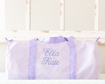 Puede incluir: Una bolsa de viaje de lona a rayas lavanda y blanca con un asa de cinta de satén lavanda. La bolsa está bordada con el nombre "Ella Kate" en una fuente cursiva.