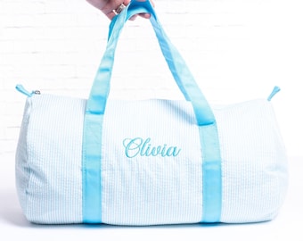Puede incluir: Una bolsa de lona azul claro y blanco a rayas con asas azules y el nombre "Olivia" bordado en la parte delantera.