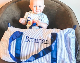 Puede incluir: Una bolsa de viaje azul y blanca a rayas con una correa azul marino y el nombre "Brennan" bordado en la parte delantera. Un niño pequeño está sentado en una silla detrás de la bolsa.